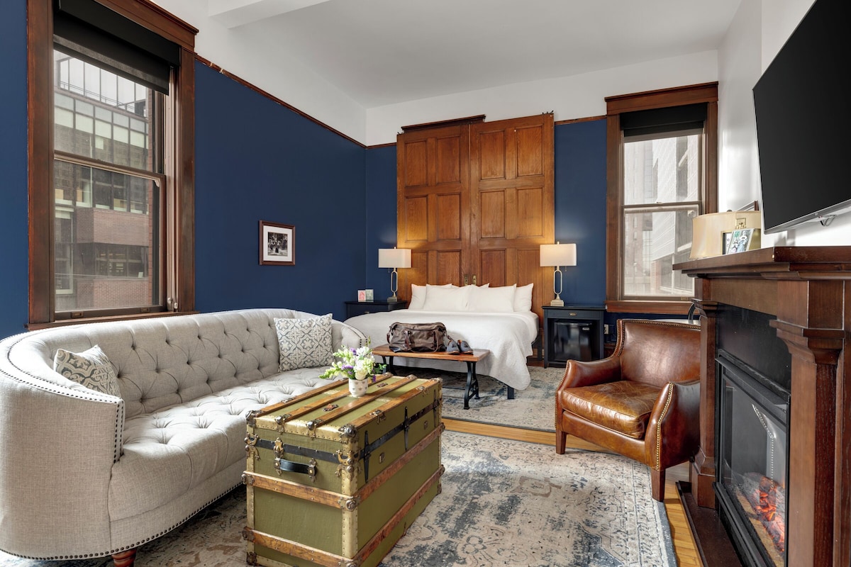 A room with a large bed, a sofa, a leather armchair, a wooden cabinet, and a fireplace.
