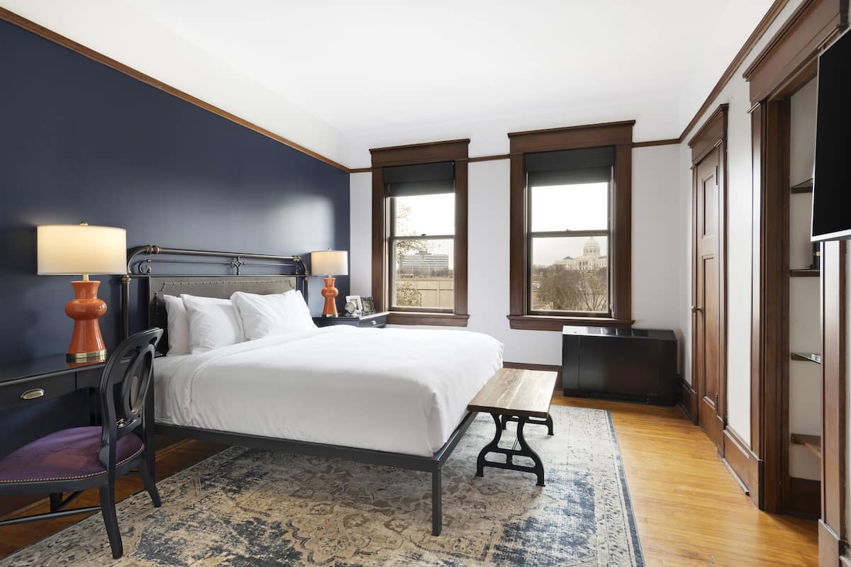 A bedroom with a bed, bedside tables, a chair, a bench, and a view of a building through windows.
