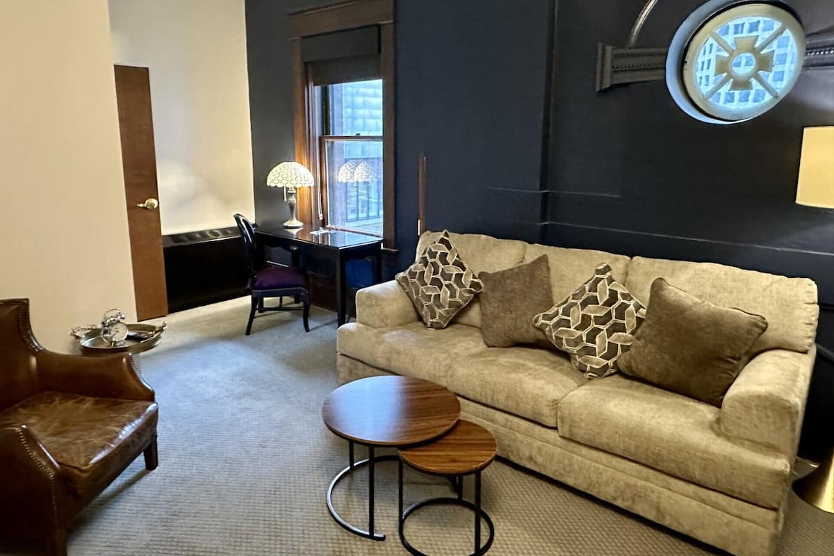 A living room with a beige sofa, a wooden coffee table, a leather armchair, a desk with a lamp, and a wall clock.