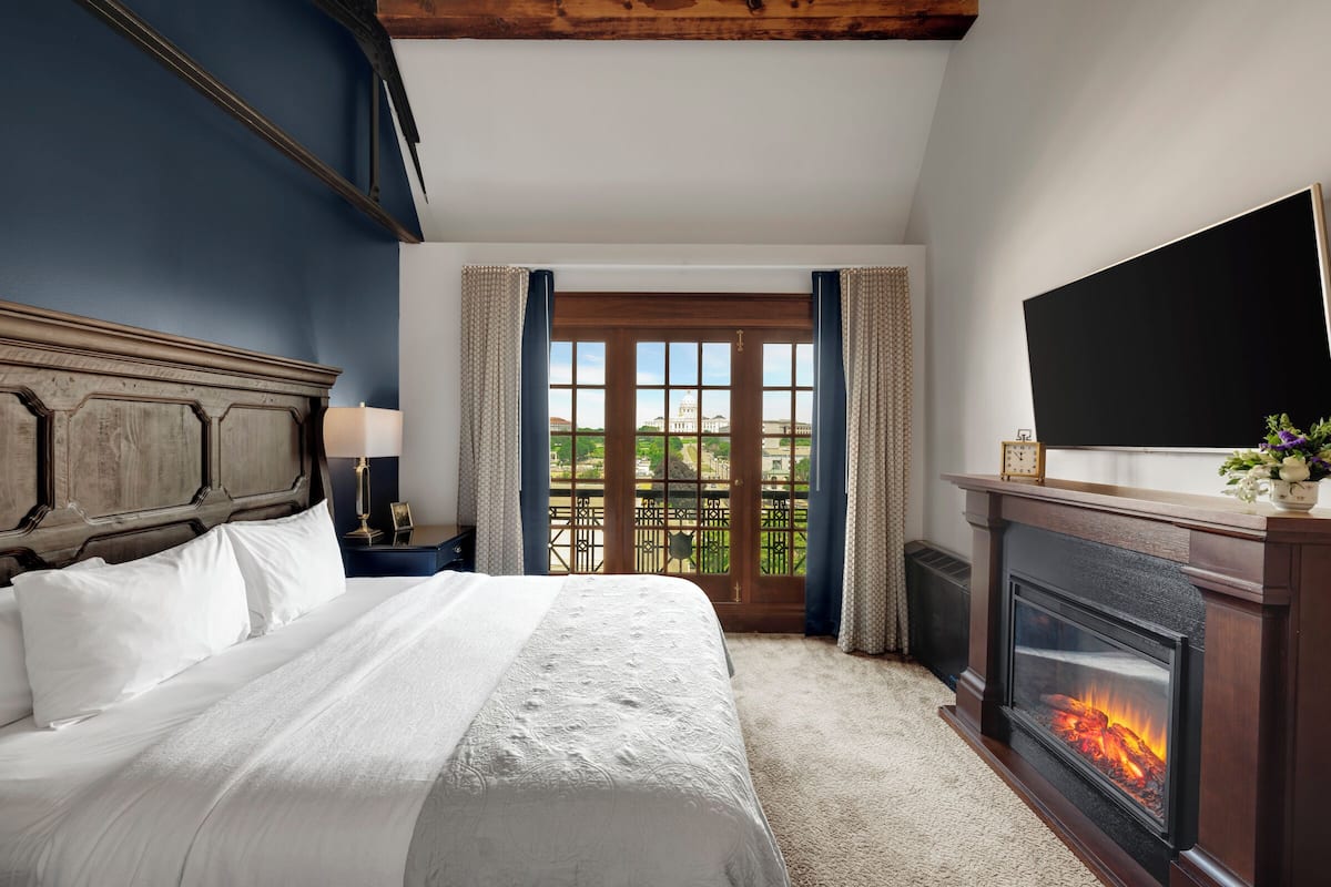 A hotel room with a large bed, a fireplace, a television, and a balcony with a view of a building.