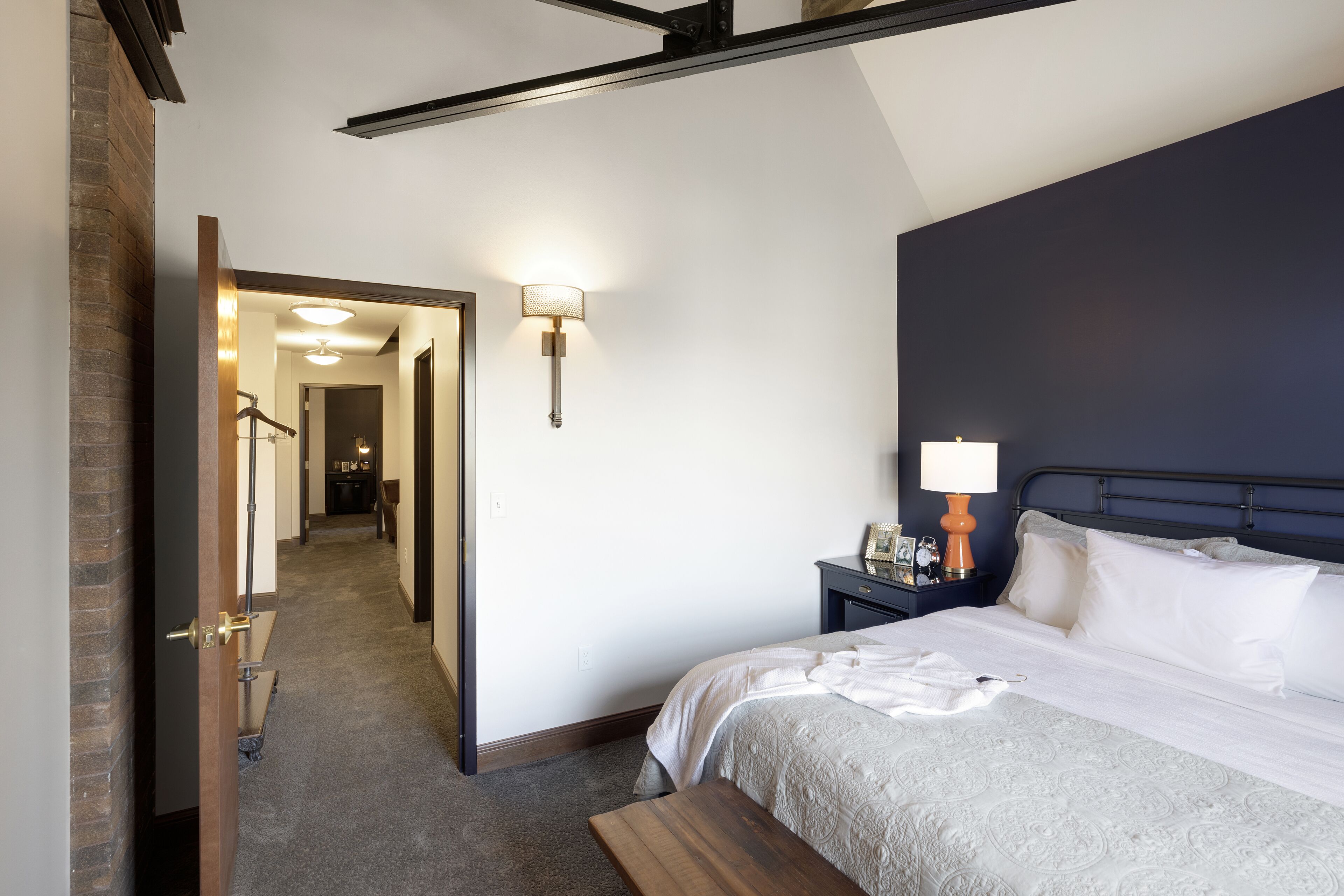 Een hotelkamer met een groot bed, een nachtkastje met een lamp, een houten bankje en een spiegel aan de muur.