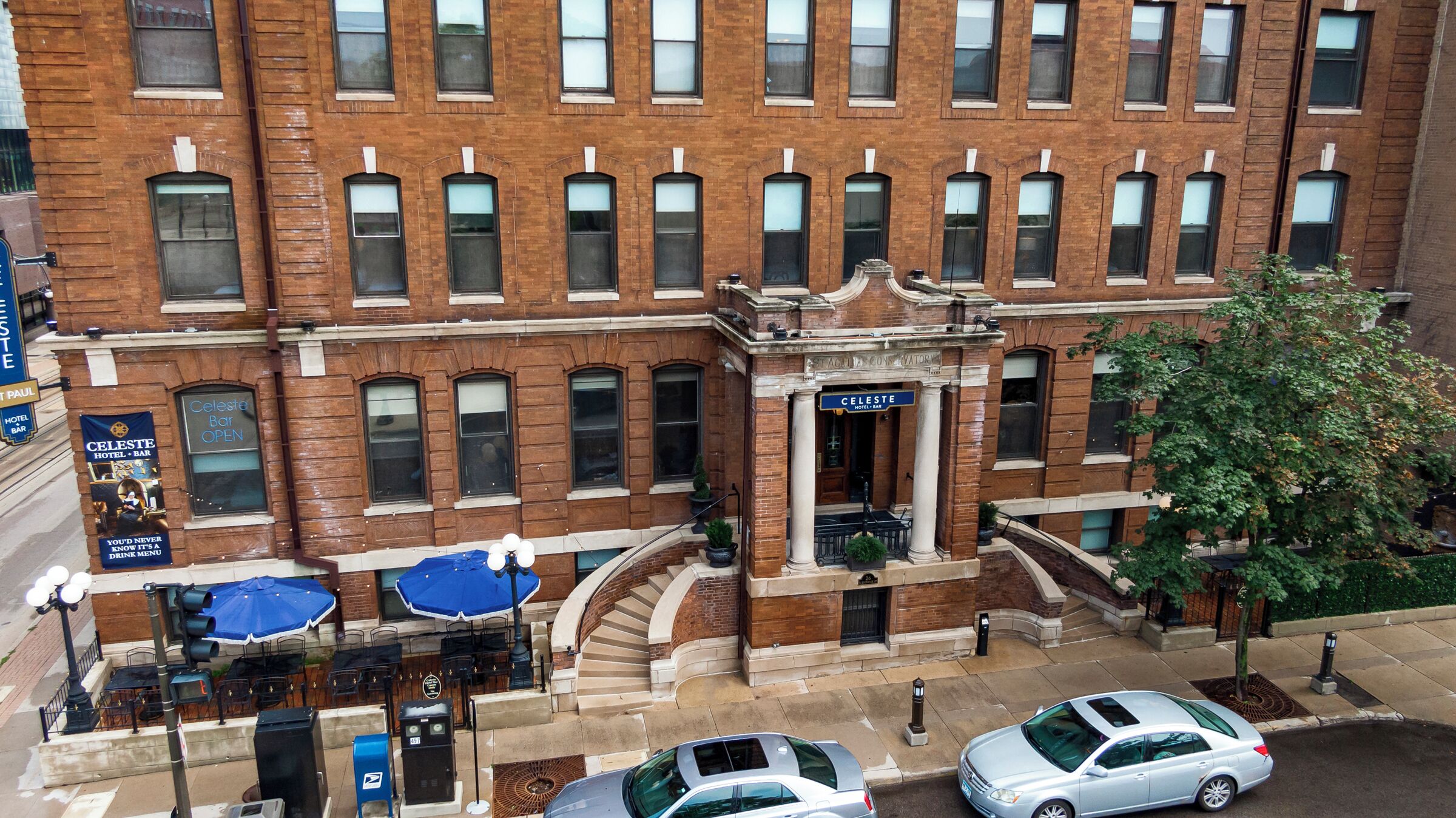 A multi-story hotel with a brick facade, a central entrance with a sign, outdoor seating with umbrellas, and a staircase leading to the entrance.