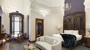 A hotel room with a large bed, a chaise lounge, a dining table with chairs, and a large window with stained glass. - Celeste of St Paul (St. Paul)