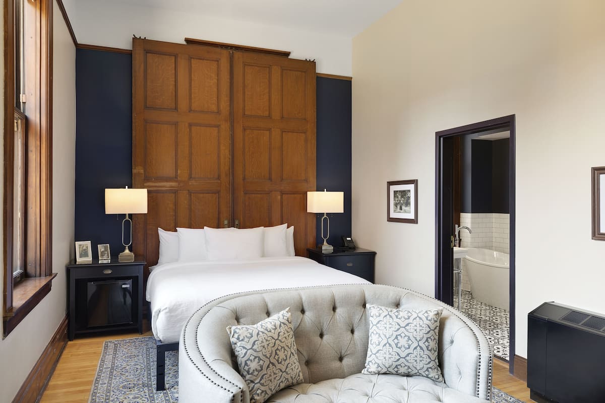 A bedroom with a large bed, two bedside tables with lamps, a grey upholstered armchair, and a wardrobe.