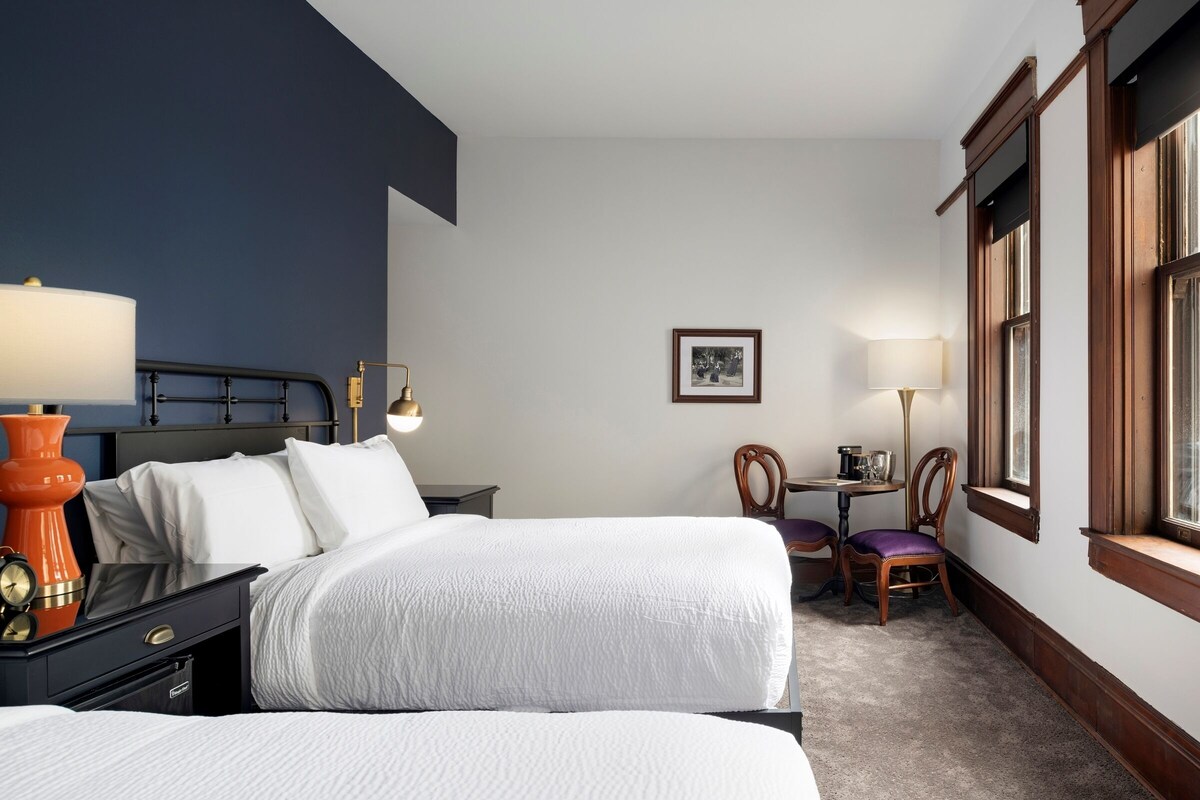 A hotel room with two beds, a nightstand with a lamp, a chair, a small table with a lamp, and a window with wooden frames.