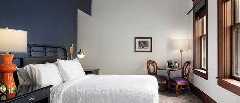 A hotel room with two beds, a nightstand with a lamp, a chair, a small table with a lamp, and a window with wooden frames.