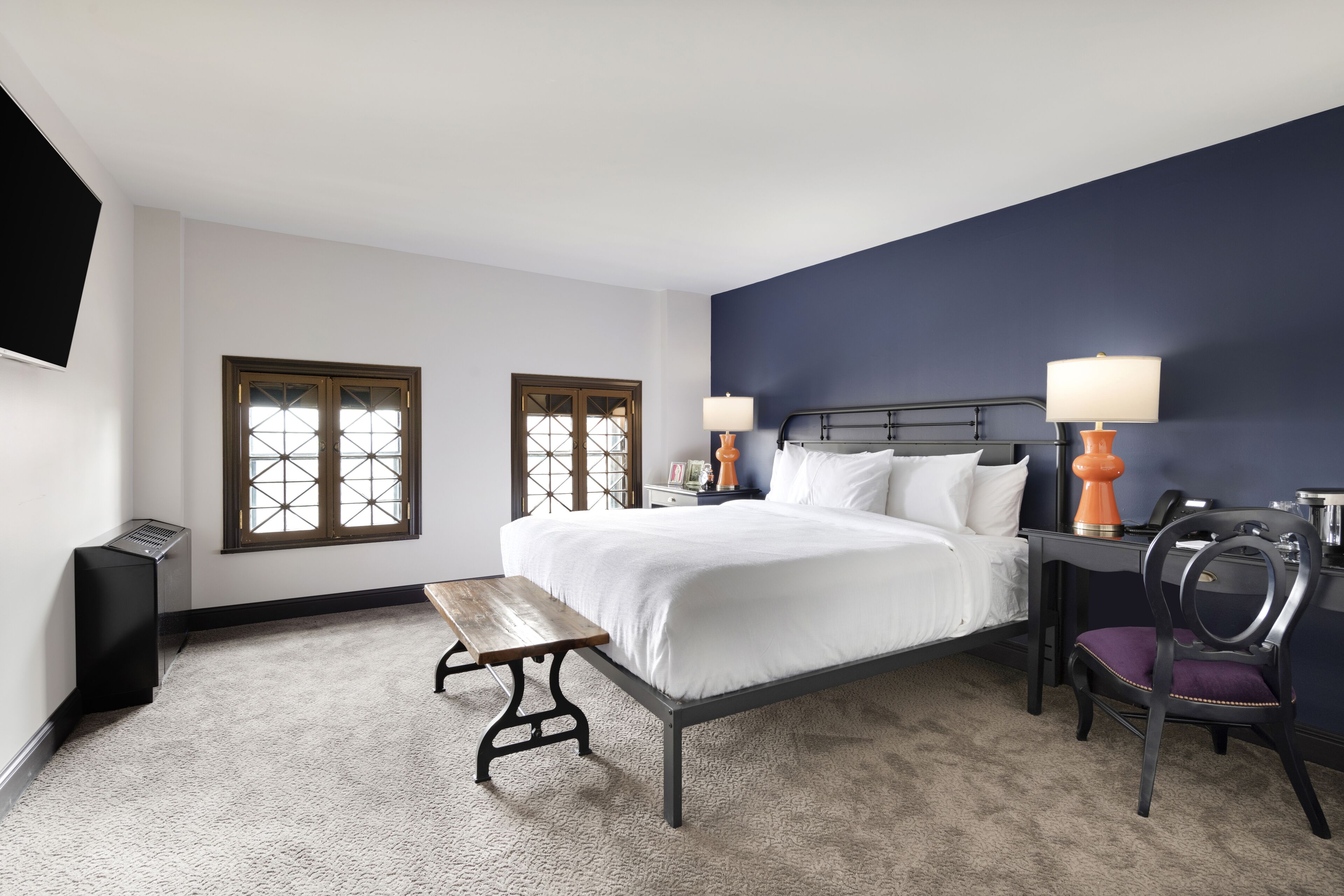 A bedroom with a bed, two bedside tables, a bench, a chair, and a TV mounted on the wall.