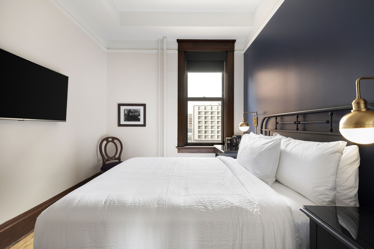 A bedroom with a large bed, a nightstand, a lamp, a framed picture, and a window with a view of buildings.