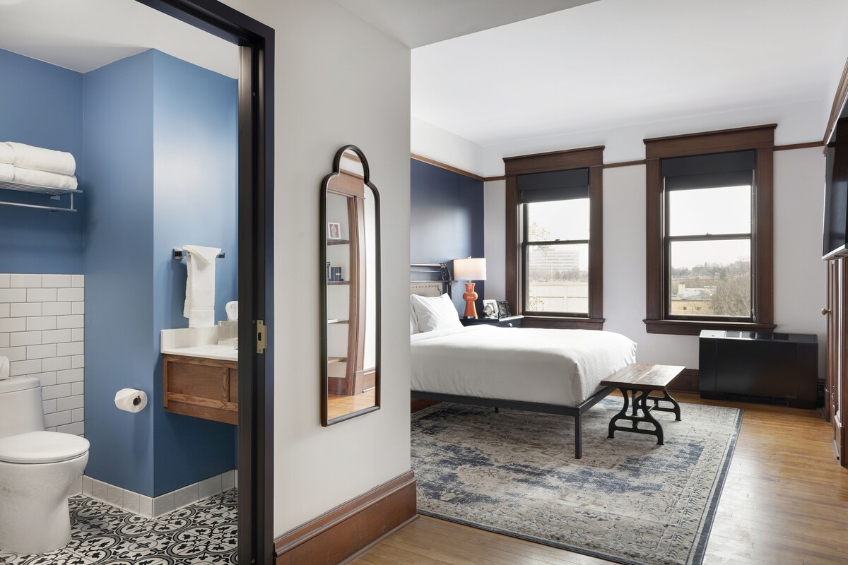 A bedroom with a large bed, a bedside table, a mirror, and a window with a view.