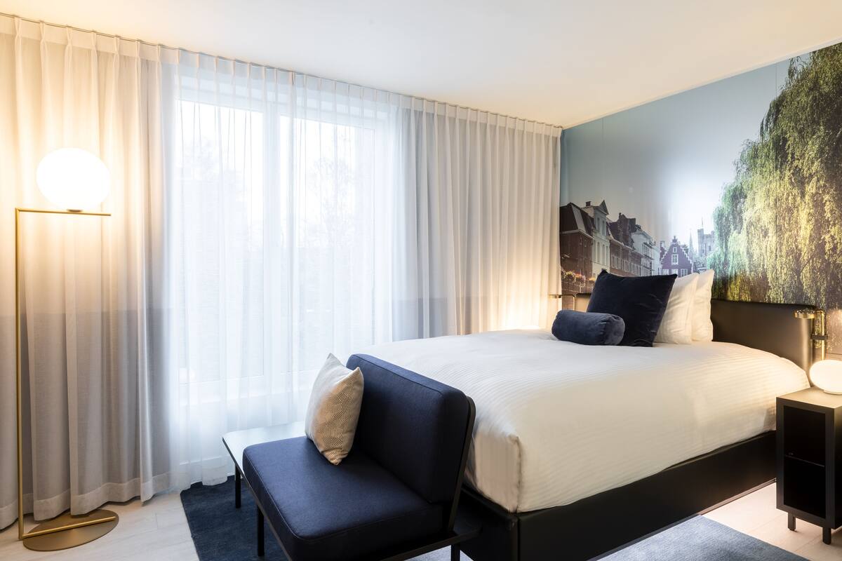Ein modernes Hotelzimmer mit einem großen Bett, einem Nachttisch, einem Sessel und Blick auf eine Stadtkulisse durch transparente Vorhänge.