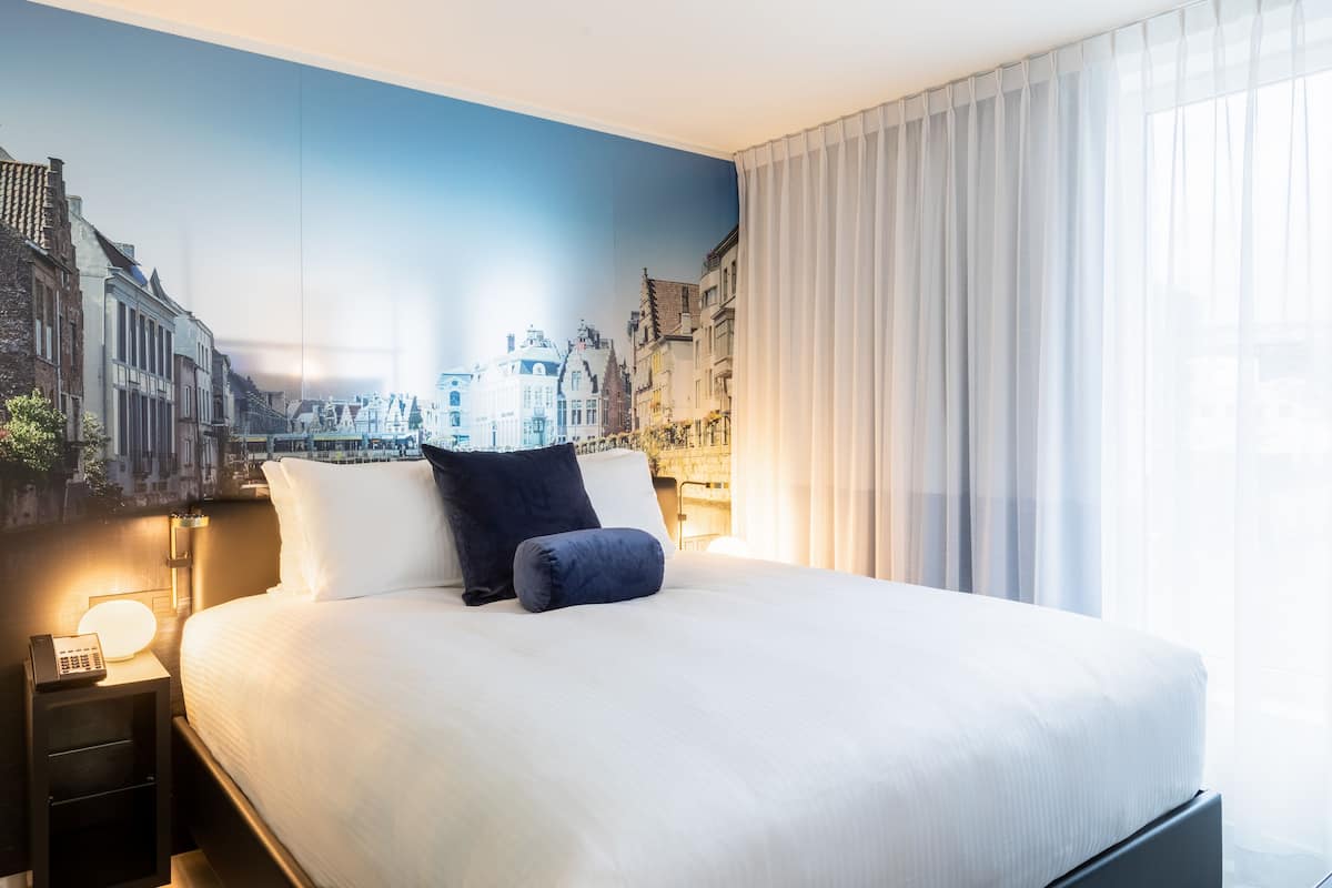 Ein Hotelzimmer mit einem großen Bett, einem Nachttisch mit Lampe und einem Stadtbild-Wandgemälde.