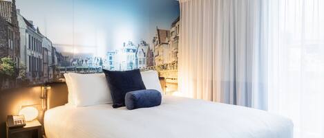 Habitación de hotel con una cama grande, una mesita de noche con lámpara y un mural de un paisaje urbano en la pared.