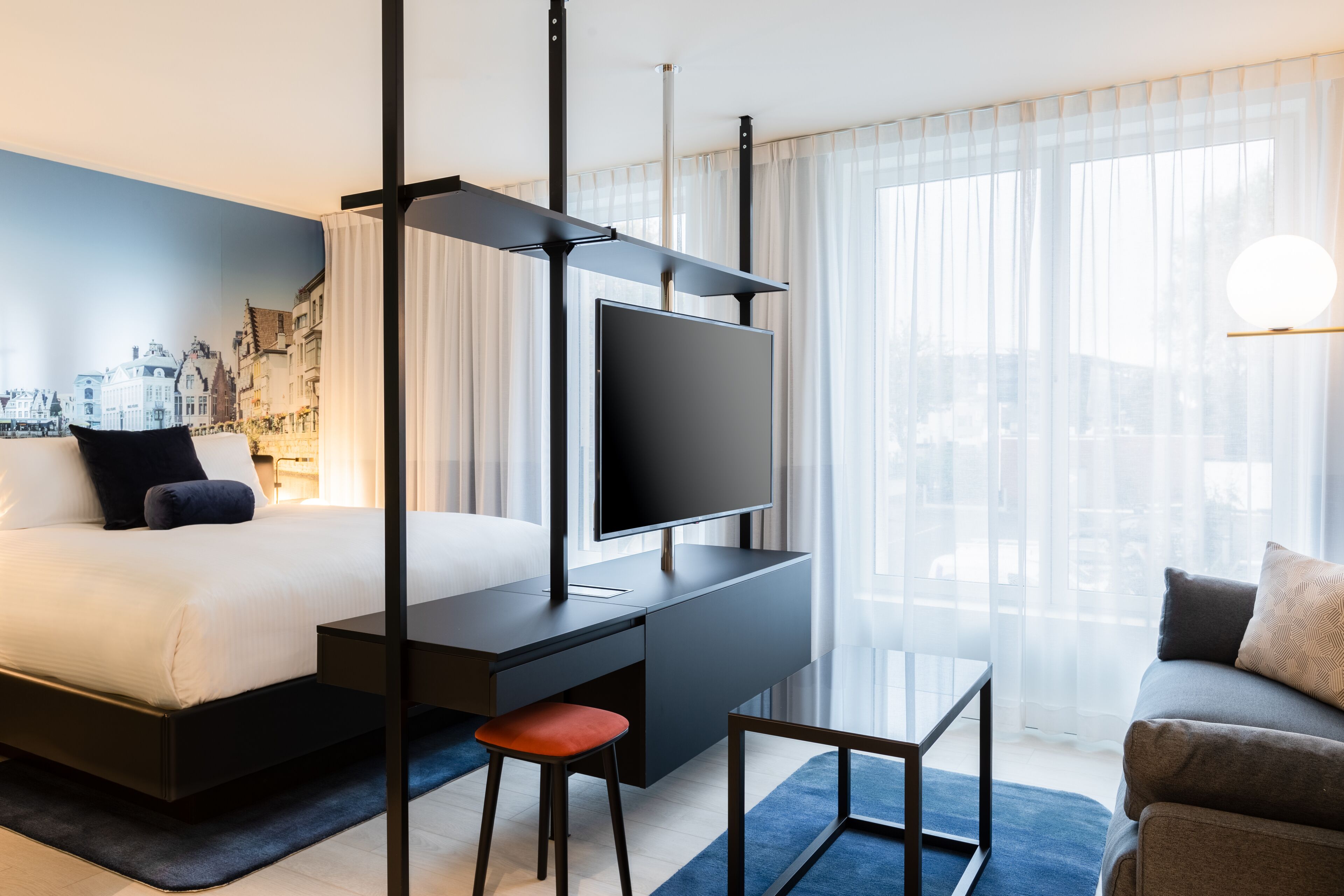 A modern hotel room with a bed, a desk with a TV, a sofa, and a cityscape mural on the wall.