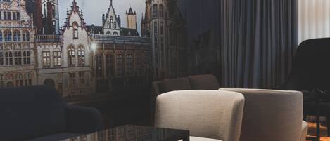 A room with a cityscape mural, a black table, two grey armchairs, and an orange carpet.