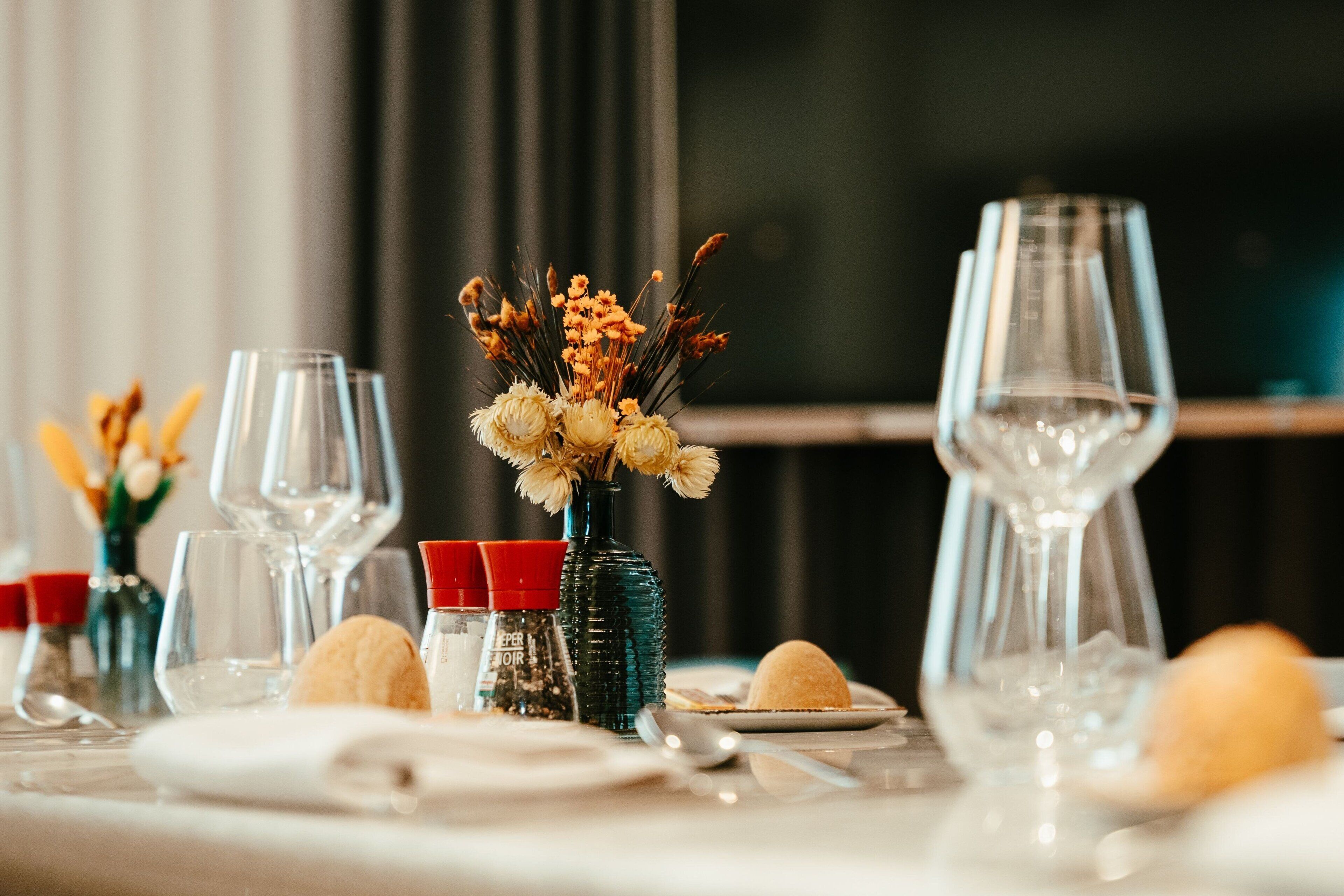 A dining table set with wine glasses, a vase with dried flowers, salt and pepper shakers, and bread.