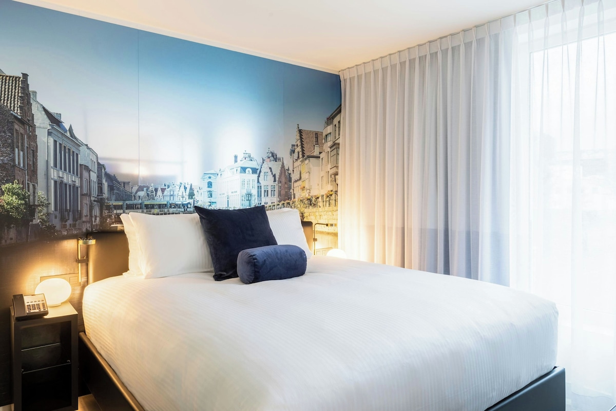 Una habitación de hotel con una cama grande, una mesita de noche, una lámpara y un mural con vistas a la ciudad.