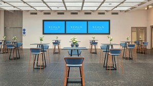 A modern event space with multiple tables and chairs, a large screen displaying 'SCALA EVENT CENTER', and a clean, minimalist design.