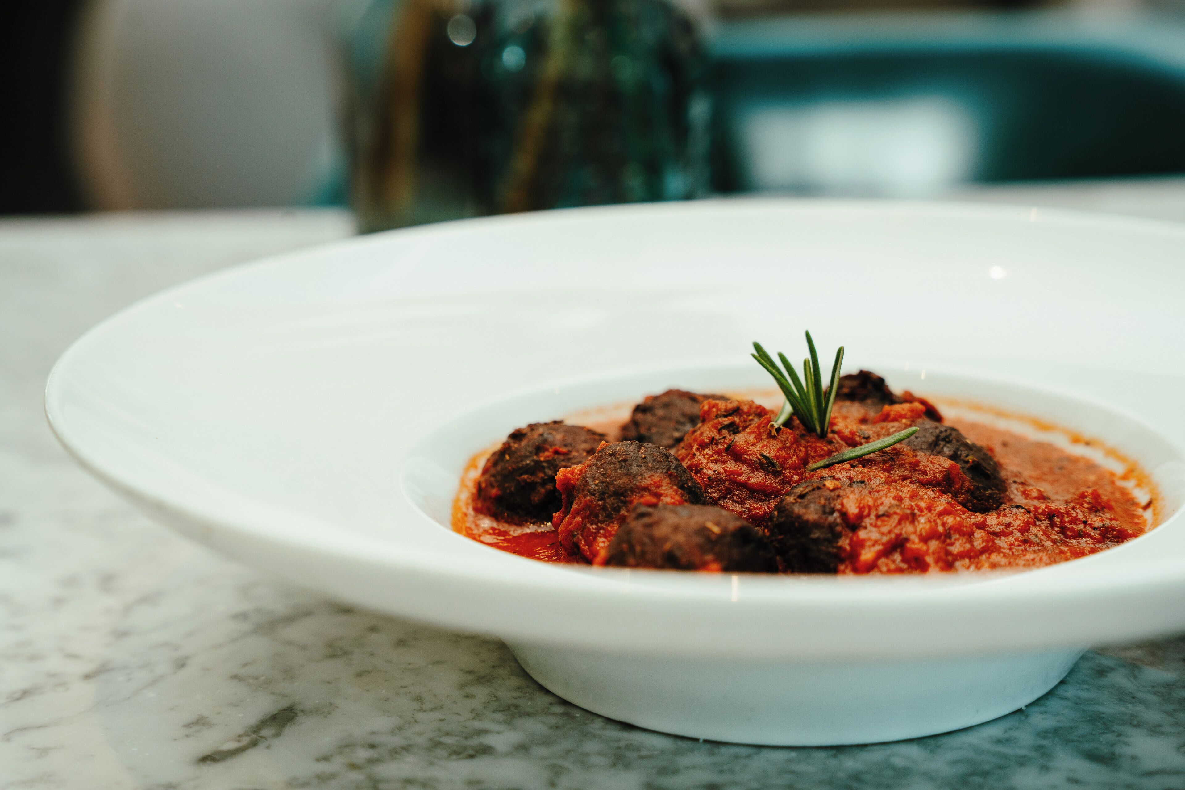 A white plate with meatballs in tomato sauce, garnished with a sprig of rosemary.