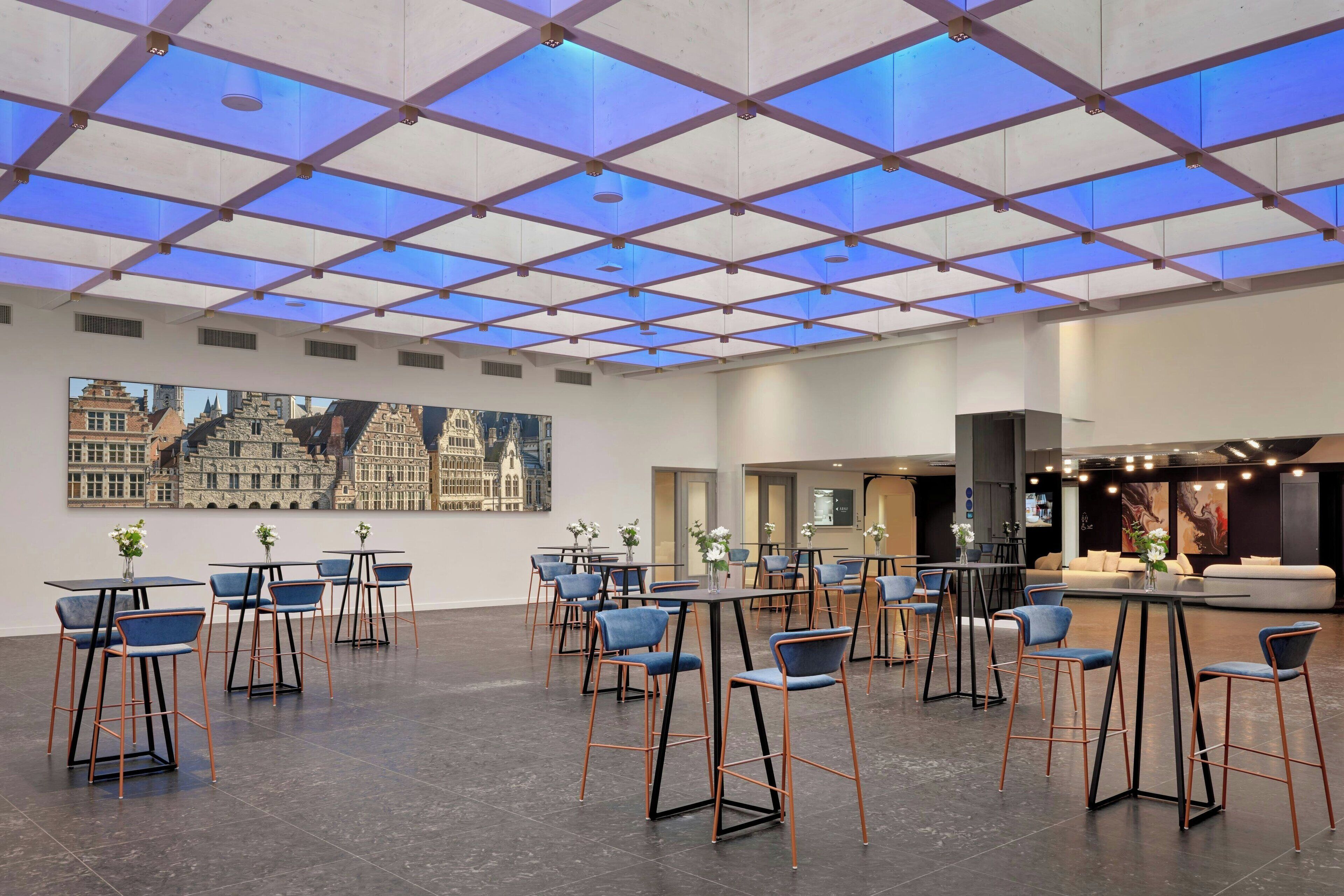 Amplio espacio interior con un diseño de techo moderno, varias mesas y sillas dispuestas para comer o reuniones, y una gran pintura de un paisaje urbano en la pared.