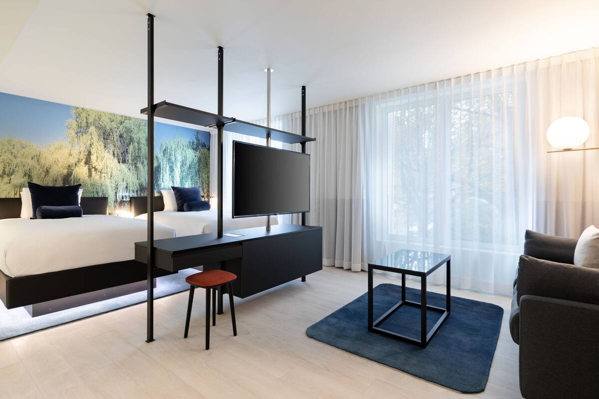 Ein modernes Hotelzimmer mit Bett, Schreibtisch, Fernseher und Blick auf Bäume durch leichte Vorhänge.