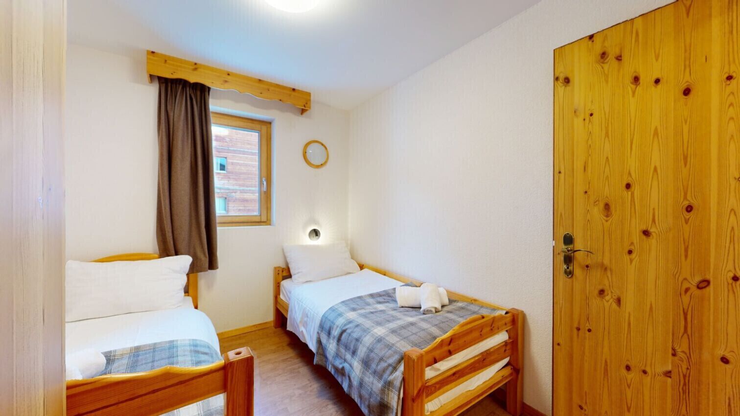 A bedroom with two single beds, a wooden door, a window with curtains, and a wall-mounted light fixture.