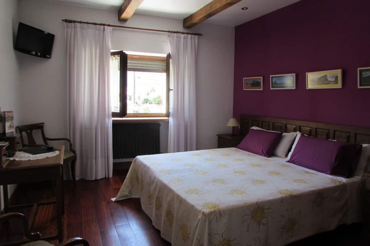 Quarto com cama, escrivaninha, cadeira, janela com cortinas e televisão na parede.