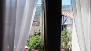 Una ventana con cortinas blancas, vista de un edificio con techos de tejas rojas, y un banco de madera afuera.