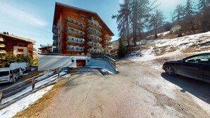 Un edificio de varios pisos con balcones, un estacionamiento con vehículos y un paisaje nevado con árboles.