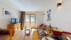 Una habitación acogedora con un sofá de madera, un televisor sobre un mueble de madera, una mesa de comedor preparada para dos personas y un balcón con vistas a un paisaje nevado.