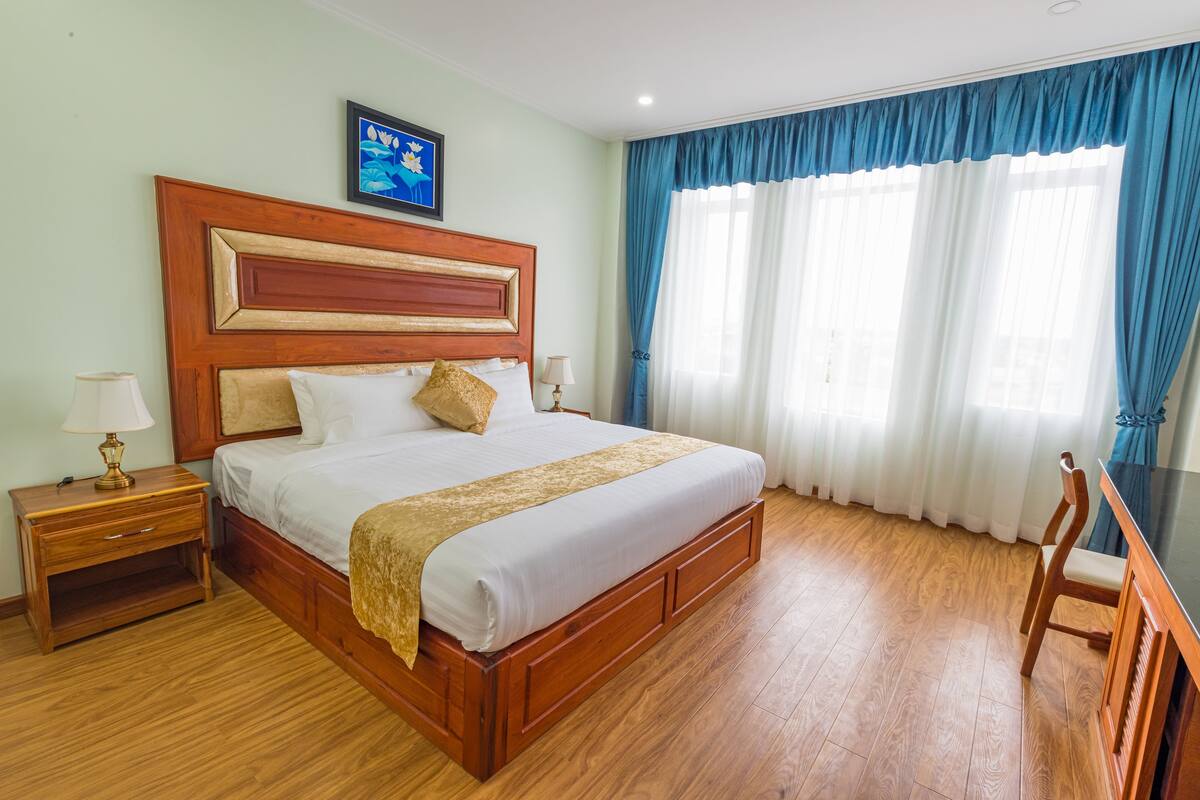 Ein Hotelzimmer mit einem hölzernen Bett, einem Nachttisch, einem Schreibtisch und einem Fenster mit blauen Vorhängen.