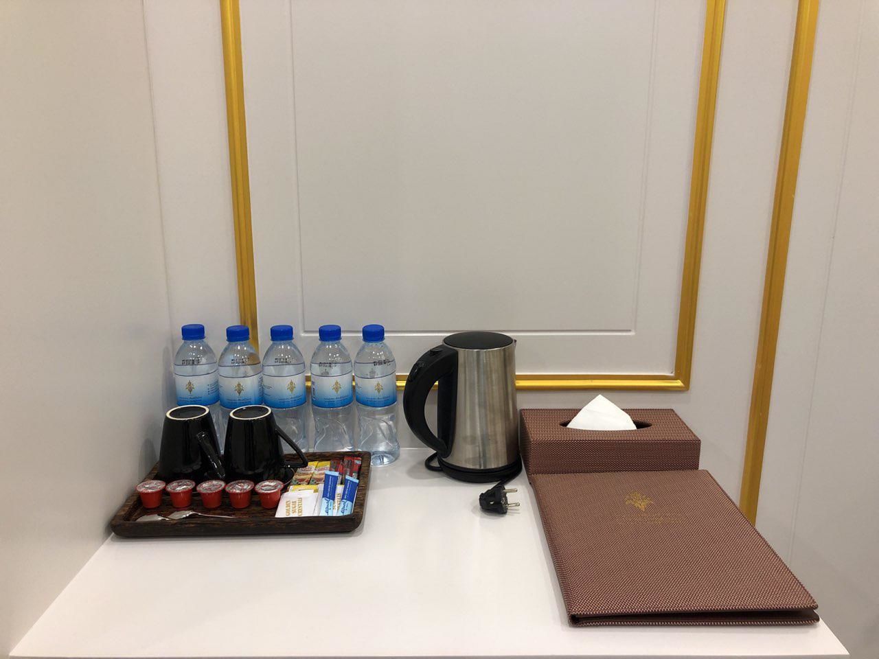 A hotel room with a white table, a brown tray with cups and bottles, a black coffee pot, a brown tissue box, and a water bottle.