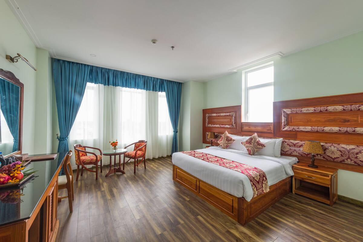 Ein Hotelzimmer mit einem hölzernen Bett, einem Schreibtisch mit Stuhl, einem kleinen Tisch mit Stuhl und einem großen Fenster mit Vorhängen.