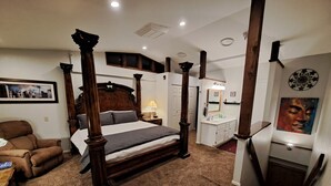Una cama con dosel de estructura de madera oscura, cabecera blanca y alfombra color beige. Hay un sillón de cuero marrón y una mecedora de madera en la habitación.
