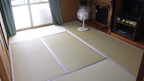 Een kamer met tatami matten, een staande ventilator, een plank met kussens en een tv-meubel met Minion beeldjes.