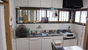 Een compacte keuken met witte kastjes, een magnetron, een spoelbak en een fornuis. Er is een raam boven de spoelbak en een klein tafeltje met papieren en een notitieboekje.