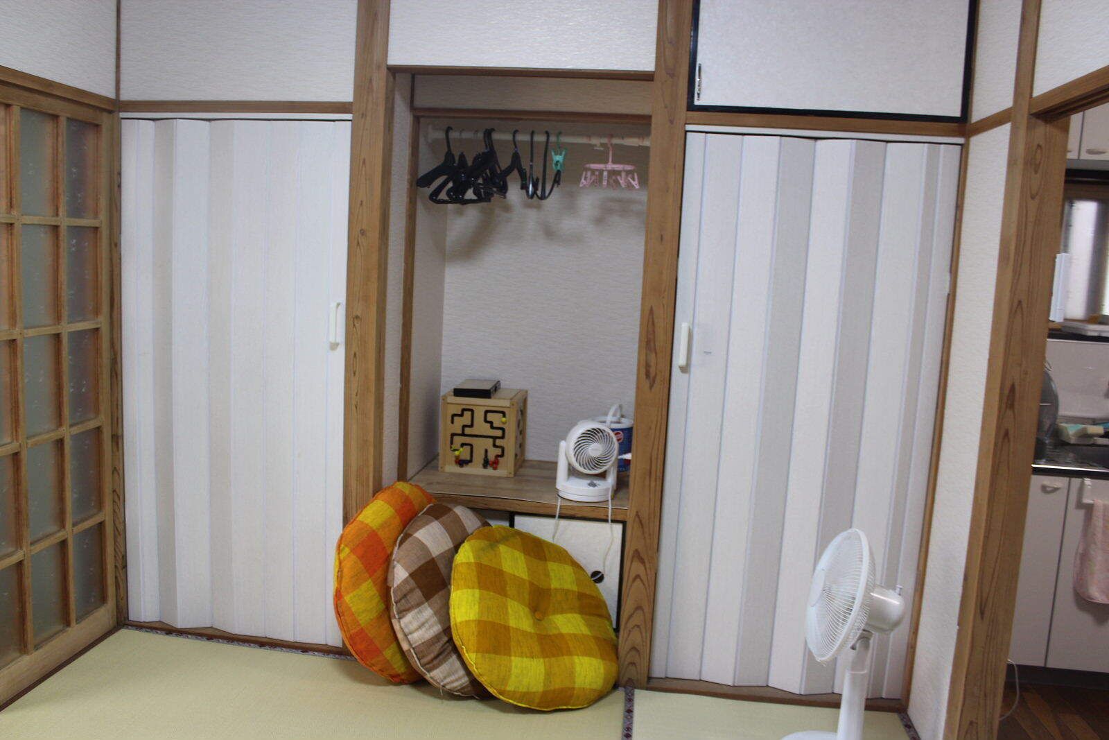 A room with a wooden wardrobe, a small shelf with a fan, and a white fan on the floor.