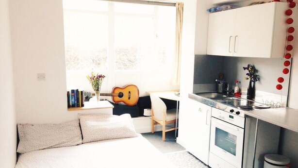 Ett litet sovrum med en säng, ett litet skrivbord, en gitarr och ett fönster.