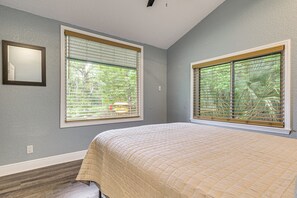 A bedroom with a bed, two windows with blinds, a wall-mounted mirror, and a door.