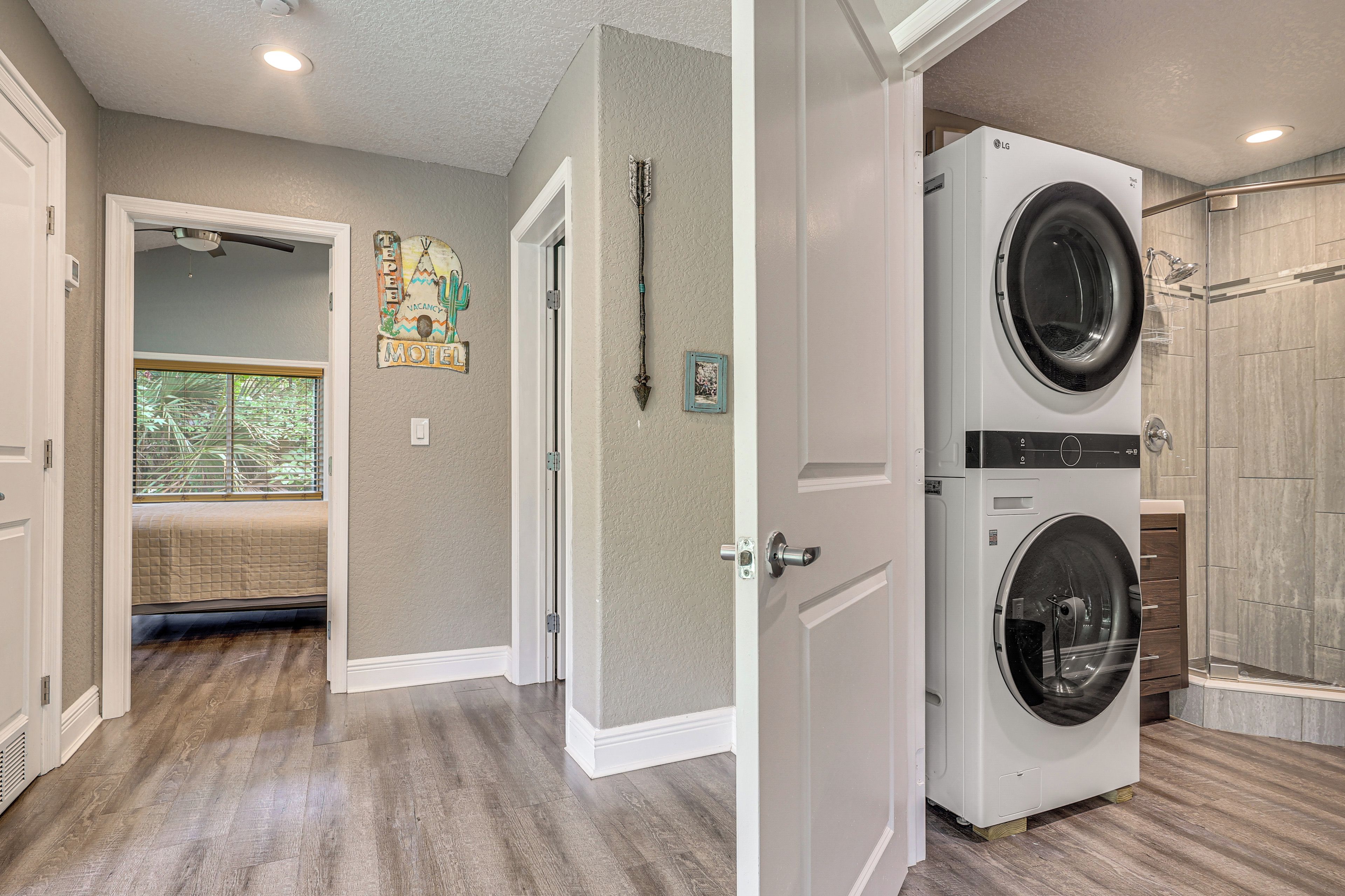 A laundry room with a front-loading washer and dryer, a shower with a glass door, and a door leading to a bedroom with a bed and a window.