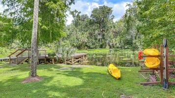 Une zone herbeuse avec une terrasse en bois, un kayak jaune et une planche de paddle, le tout posé sur un support en bois.