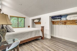 A bedroom with a bed, a bedside table with a lamp, a window with blinds, a shelf with folded blankets, and a wooden floor.