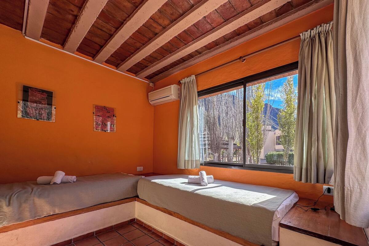 Una habitación con dos camas, un equipo de aire acondicionado, una ventana con cortinas y vistas a árboles y edificios.