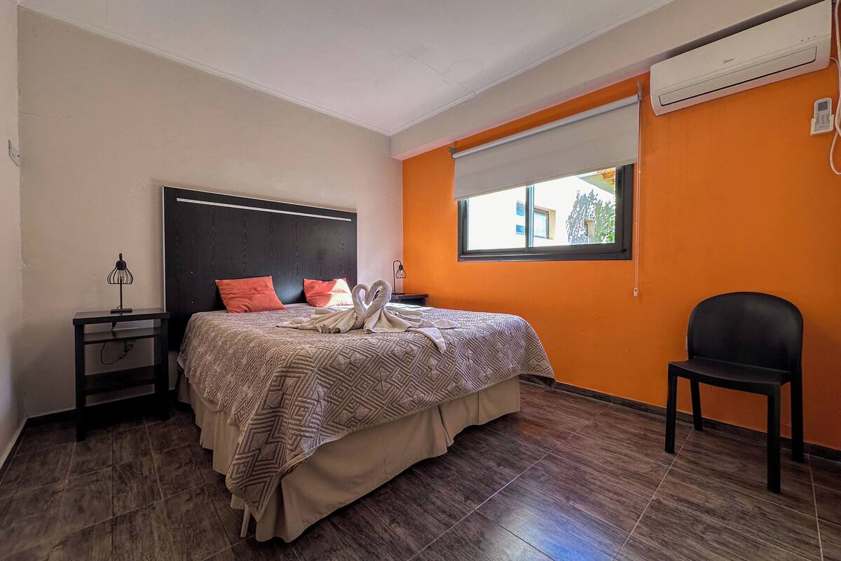 Habitación de hotel con cama, mesita de noche, silla, unidad de aire acondicionado y ventana con vista a edificios.
