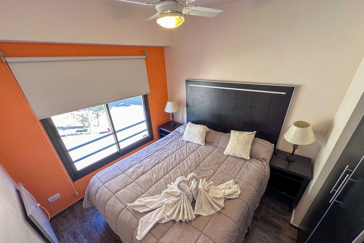 Un dormitorio con una cama grande, un ventilador de techo, una ventana con persianas y una mesita de noche con lámpara.
