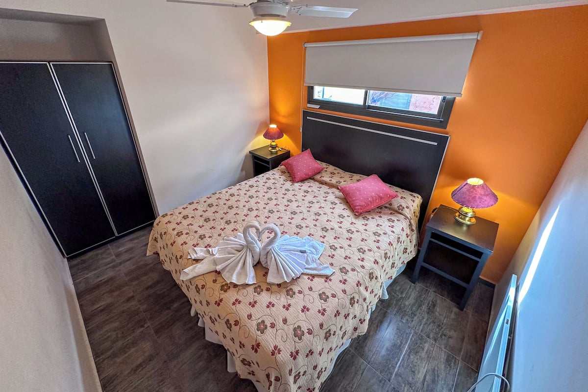 Un dormitorio con una cama, un ventilador de techo, una mesita de noche con lámpara, un armario y una ventana con persianas.