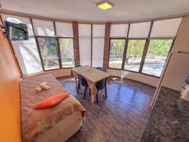 Quarto com cama, mesa, cadeiras, janelas amplas com cortinas e uma luminária no teto.