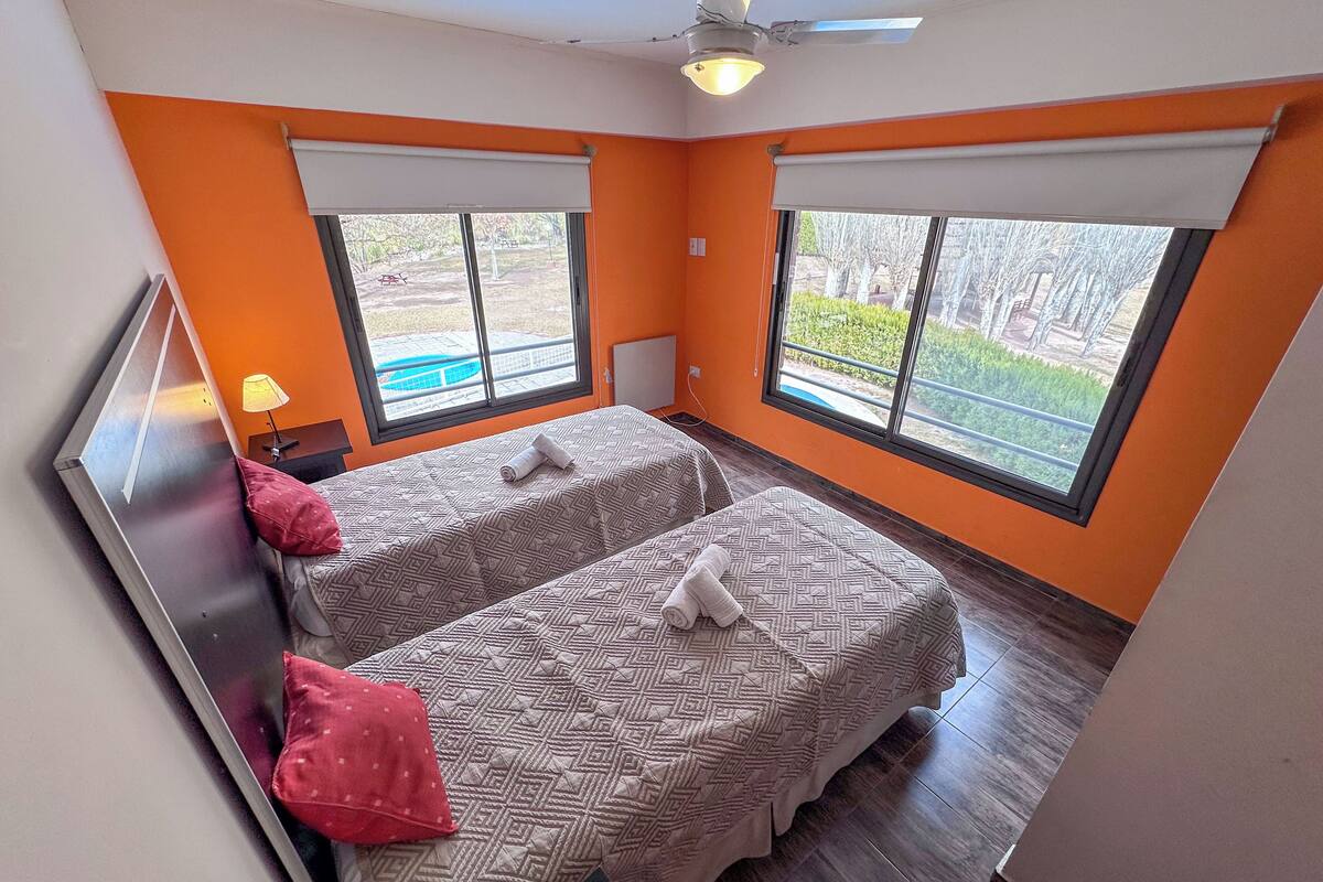 Habitación con dos camas, paredes anaranjadas, ventanas amplias, ventilador de techo y vistas al exterior.