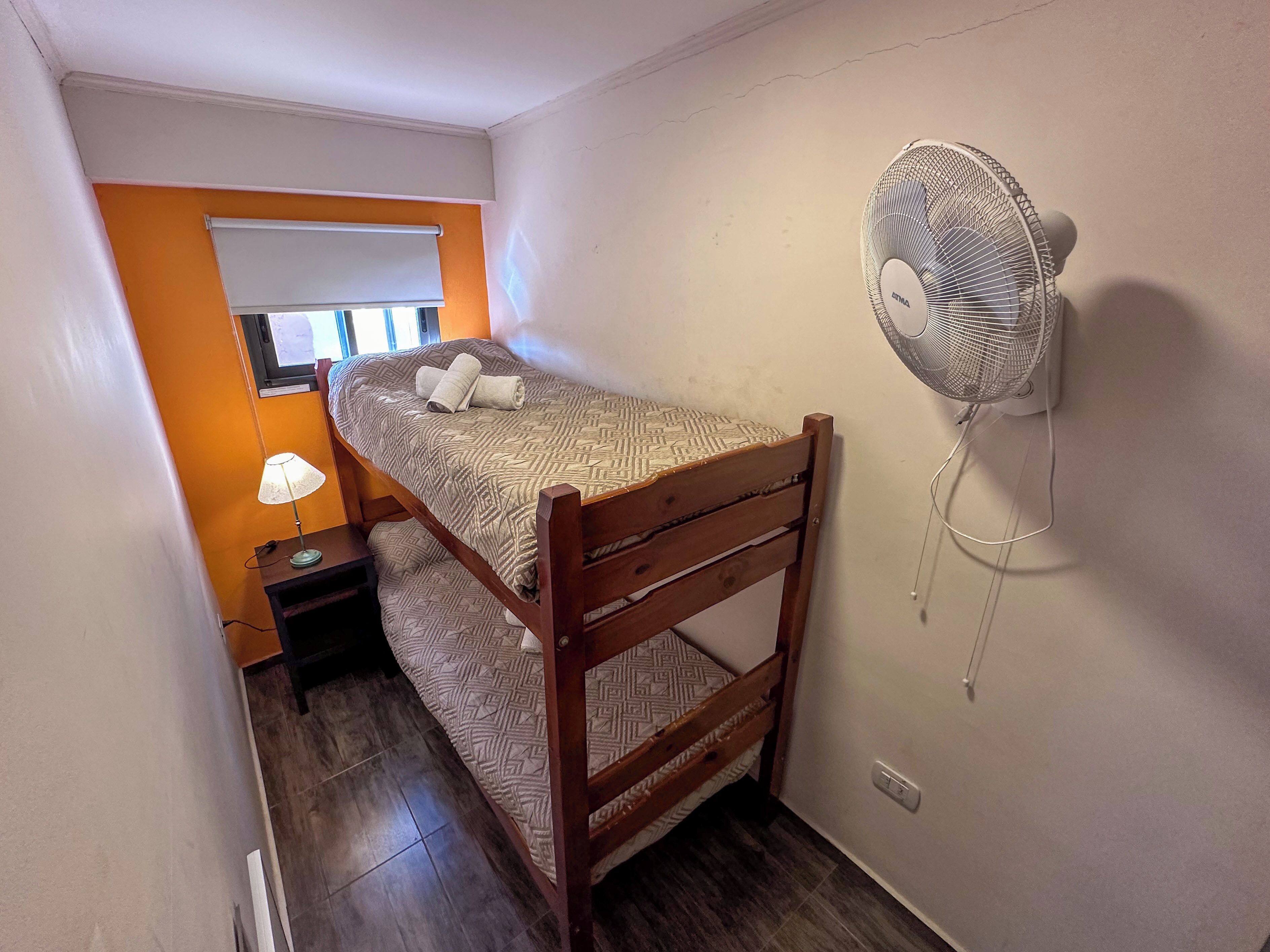 Una habitación pequeña con cama individual, estructura de madera, ventilador de pared, mesita de noche con lámpara y ventana con persiana blanca.