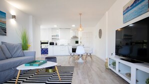 Una sala moderna con un sofá, una mesa de centro y un mueble para televisión. Se ve una cocina al fondo con armarios blancos y una zona de comedor con sillas.