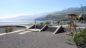 Una playa con una estructura de madera, escalones que conducen a una zona más elevada, y una vista del mar y montañas al fondo.